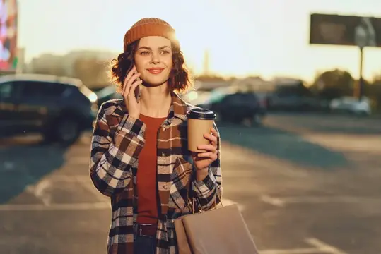 Улыбающаяся девушка в шапке говорит по телефону с кофе на парковке