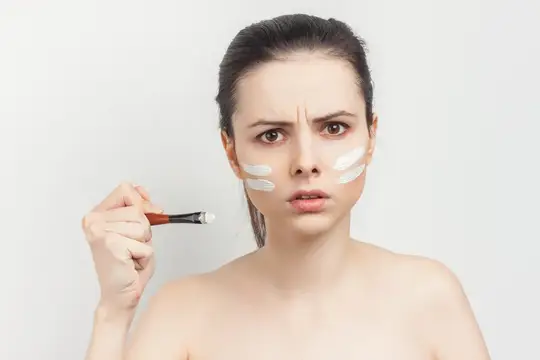 Брюнетка с обнаженными плечами наносит макияж на лицо, кадрированный вид