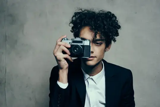 Кудрявый парень в пиджаке фотографирует на плёночный фотоаппарат