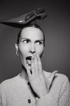 Светлокожая женщина-европеоид с короткими волосами позирует удивлённо с туфлей на голове, в серьгах и вязаном свитере