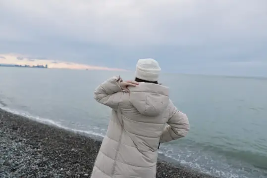 Девушка в белом пуховике стоит спиной к камере на галечном берегу