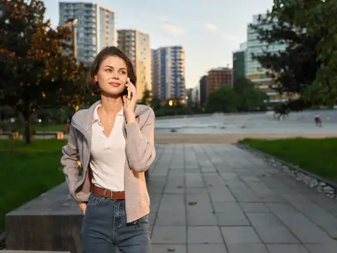 Женщина стоит в городском парке, пользуясь мобильным телефоном, на заднем плане высокие здания и зеленые насаждения