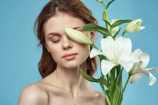 Молодая женщина с закрытыми глазами и белыми лилиями у лица на голубом фоне