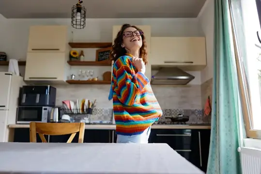 Кудрявая женщина в очках смеётся на кухне в разноцветном свитере