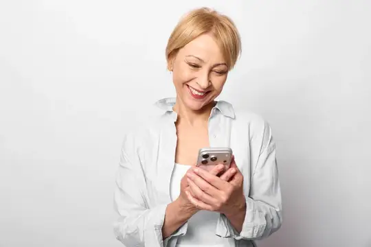 Улыбающаяся блондинка в белой рубашке смотрит в смартфон на светлом фоне
