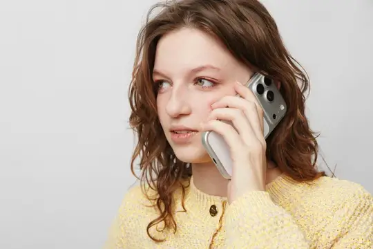 Женщина с вьющимися волосами в желтом вязаном свитере держит смартфон у уха, глядя в сторону