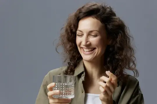 Улыбающаяся кудрявая женщина держит стакан воды на сером фоне
