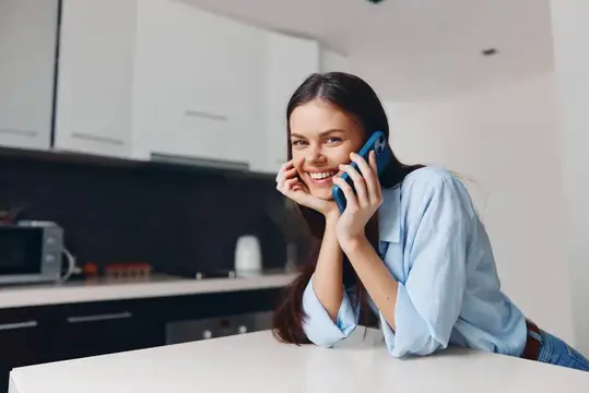 Улыбающаяся темноволосая девушка говорит по телефону на светлой кухне