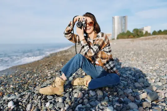 Улыбающаяся девушка в клетчатой рубашке фотографирует на каменистом берегу