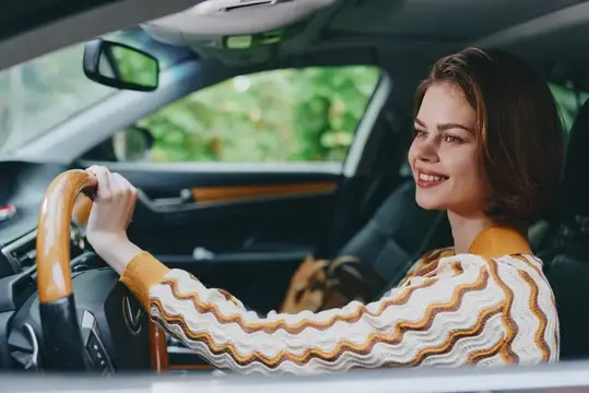 Улыбающаяся темноволосая женщина за рулём автомобиля смотрит в боковое окно