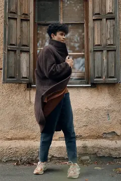 Модная женщина в коричневом пальто и джинсах стоит на фоне окна старого здания с деревянными ставнями