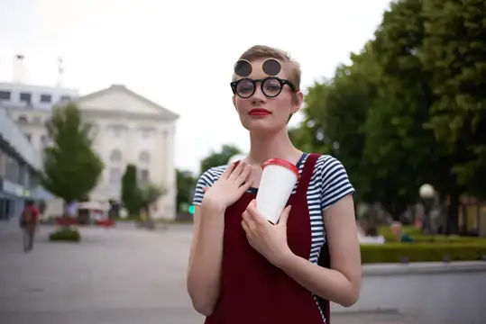 Женщина с короткой стрижкой стоит на улице в солнцезащитных очках с напитком в руке