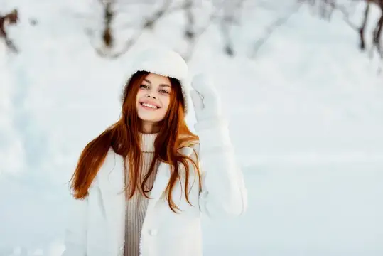 Рыжеволосая девушка в белой шапке улыбается зимой в снежном лесу