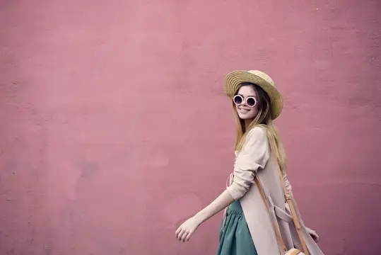 Улыбающаяся девушка в соломенной шляпе и круглых очках на фоне розовой стены