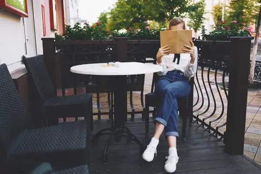 Девушка в джинсах читает книгу за столиком летнего кафе