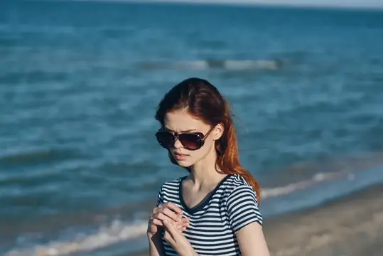 Женщина в солнечных очках стоит на пляже у моря, на заднем плане — природный пейзаж