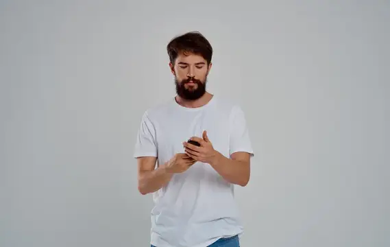 Бородатый мужчина в белой футболке смотрит в смартфон на сером фоне