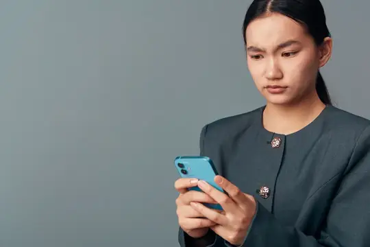 Азиатская девушка в сером пиджаке сосредоточенно смотрит в смартфон