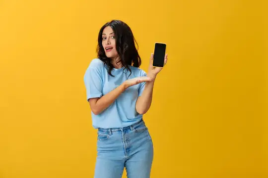 Удивлённая темноволосая девушка показывает на смартфон на жёлтом фоне