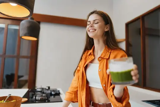 Улыбающаяся девушка с закрытыми глазами держит стакан зелёного смузи на кухне