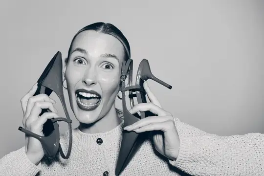 Удивлённая женщина с туфлями возле лица на сером фоне