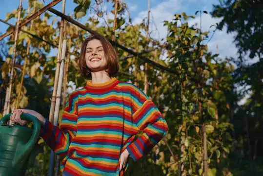 Улыбающаяся девушка в радужном свитере с лейкой в саду