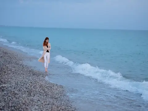 Девушка в бежевом пальто идёт босиком по кромке воды на галечном пляже