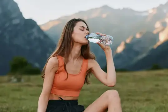 Молодая женщина в спортивном топе пьёт воду из бутылки на фоне гор
