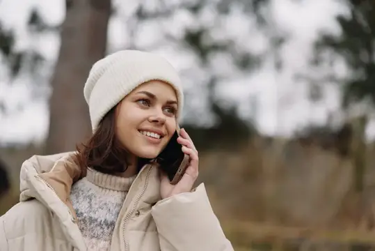 Уличный портрет улыбающейся женщины в шапке-бини, зимнем пальто и с телефоном, наслаждающейся разговором в живописном парке, передающий повседневное общение и расслабленное настроение