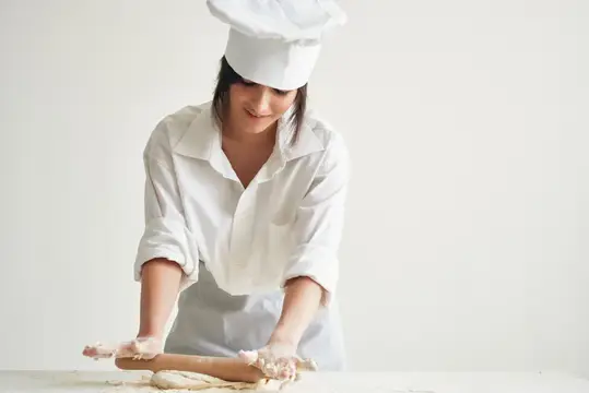 Женщина в поварской форме раскатывает тесто для мучных изделий, занимаясь домашней работой