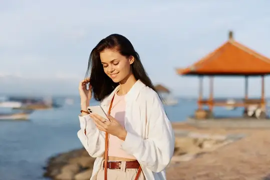 Женщина-блогер и фрилансер улыбается с телефоном в руке на отдыхе, гуляя по пляжу у океана на Бали