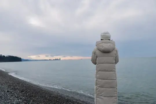 Человек в длинном пуховике смотрит на море в пасмурный день