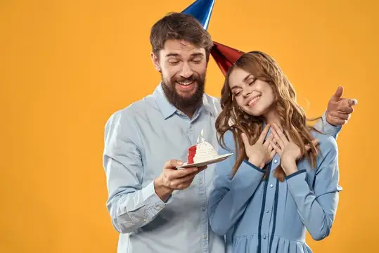 Бородатый мужчина с тортом поздравляет улыбчивую девушку в праздничных колпаках