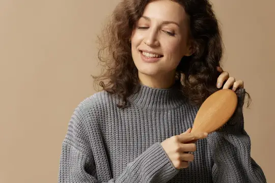 Улыбающаяся кудрявая девушка расчёсывает волосы деревянной щёткой