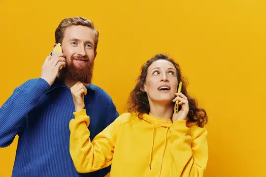 Женщина и мужчина, жизнерадостная пара, держат в руках телефоны и разговаривают по мобильному, криво улыбаясь, на жёлтом фоне