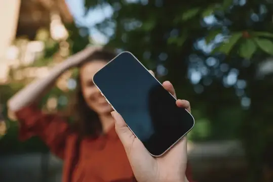 Улыбающаяся девушка держит смартфон с тёмным экраном на фоне зелени