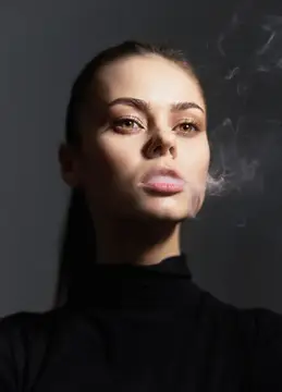 Молодая женщина с серьёзным выражением лица, одетая в чёрную водолазку, выпускает дым