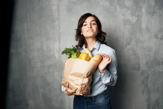 Улыбающаяся брюнетка держит бумажный пакет с продуктами у серой стены