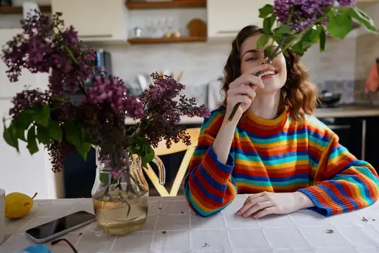 Улыбающаяся девушка в радужном свитере нюхает ветку сирени на кухне