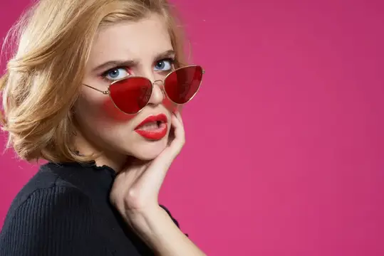 Симпатичная женщина в темных очках, ярком макияже и черной куртке крупным планом на розовом фоне