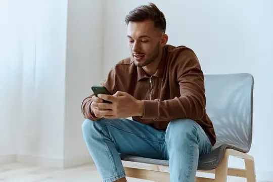 Молодой мужчина смотрит в смартфон, сидя на стуле у окна