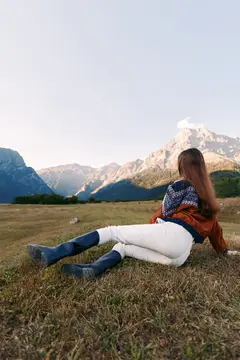 Женщина в свитере и сапогах лежит в лугу, любуясь горами и пейзажем, погруженная в природу и отдых