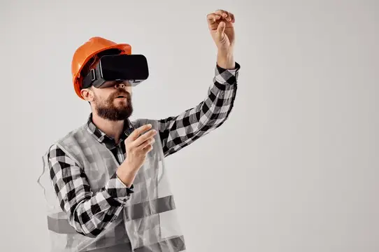 Бородатый мужчина в каске и VR-очках жестикулирует на сером фоне