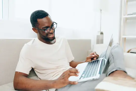 Улыбающийся темнокожий мужчина в очках работает за ноутбуком на диване