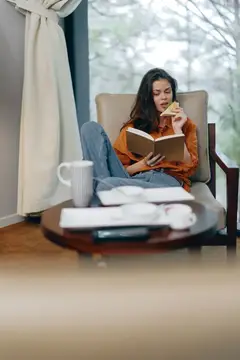 Уютно одетая женщина в повседневной одежде наслаждается чтением книги и лакомится печеньем в тёплой домашней атмосфере, куда через окно проникает естественный свет