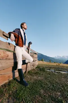 Молодой человек в кожаном пиджаке опирается на деревянную изгородь в горах