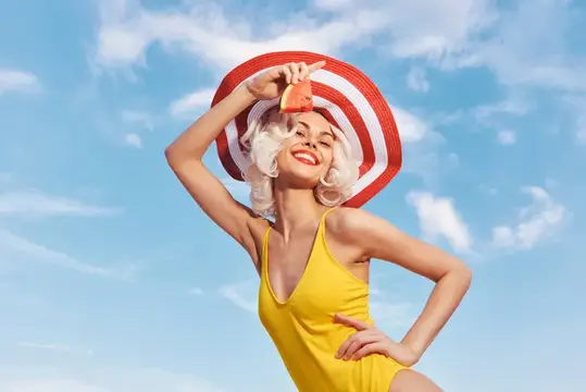 Жизнерадостная женщина позирует с улыбкой, одетая в жёлтый купальник и красно-белую полосатую шляпу, на фоне ярко-голубого неба