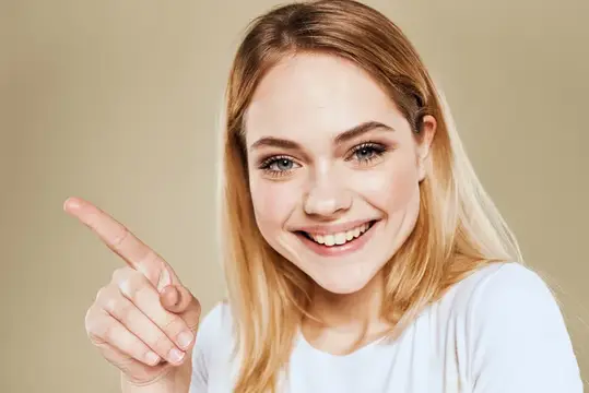 Улыбающаяся блондинка указывает пальцем в сторону на бежевом фоне