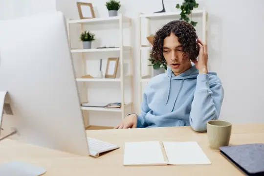 Кудрявый парень в голубом худи работает за компьютером в светлом офисе