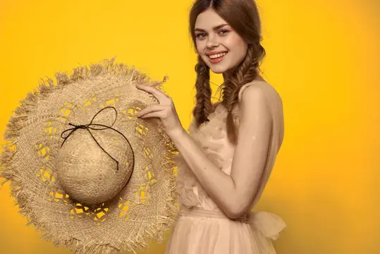 Улыбчивая девушка с косичками держит плетёную шляпу на жёлтом фоне